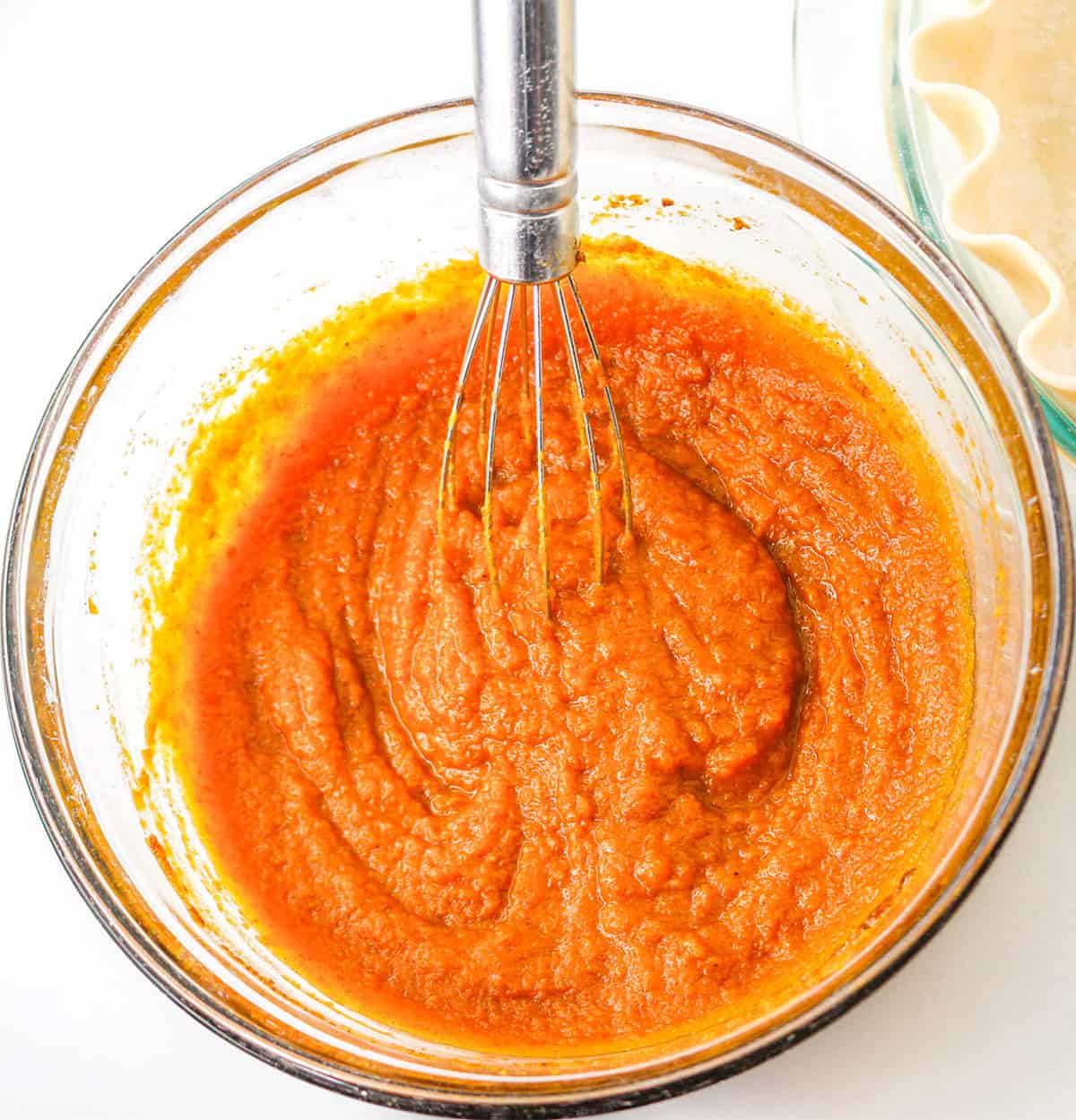 A glass bowl of orange pumpkin pie filling being whisked, with a pie crust in a separate dish visible in the upper right corner—perfect for creating a classic pumpkin pie or adding a twist with pecan pie flavors.