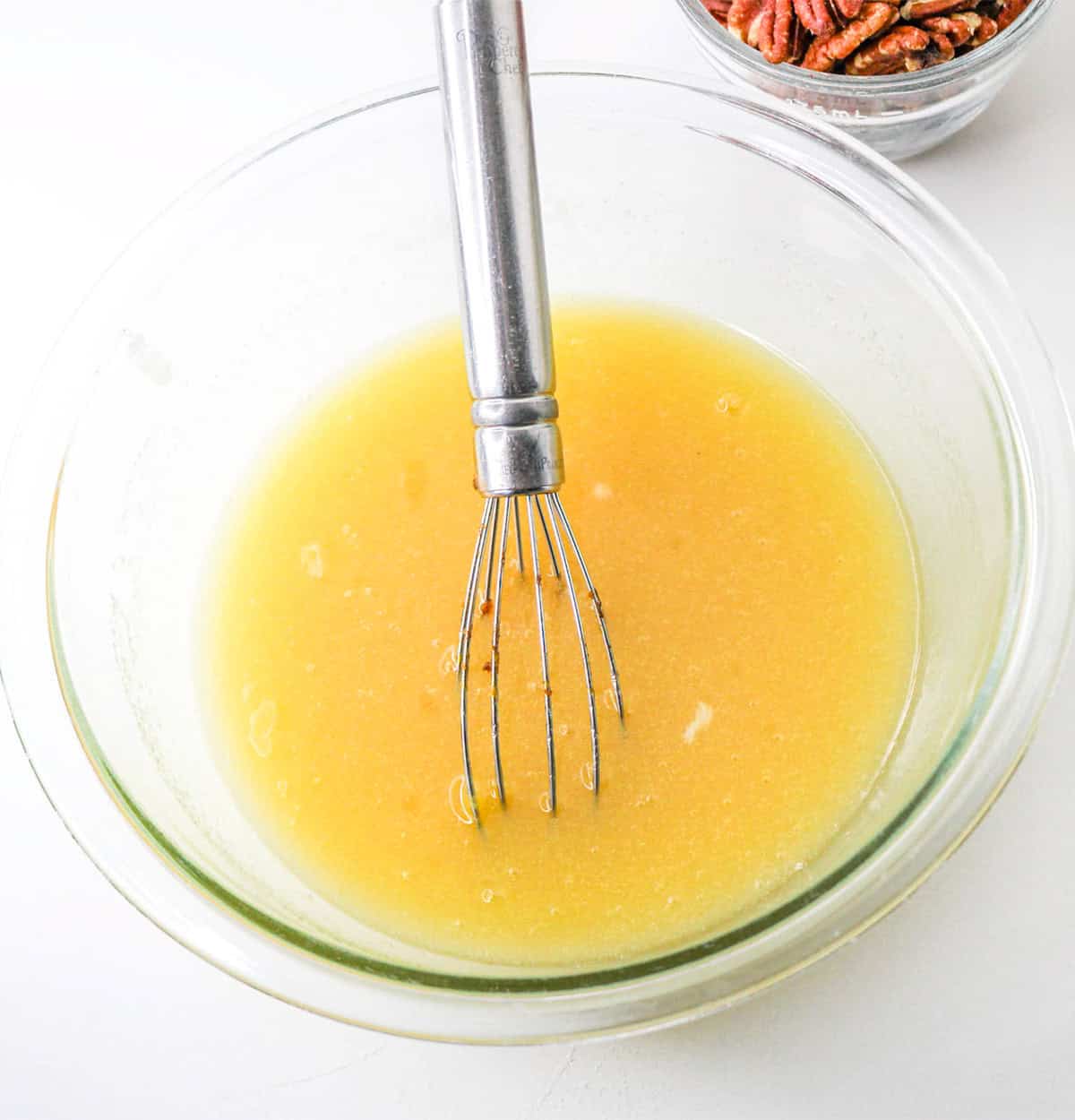 A metal whisk in a glass bowl with yellow liquid mixture, perfect for making pumpkin pie, with a bowl of pecans in the background on a white surface.