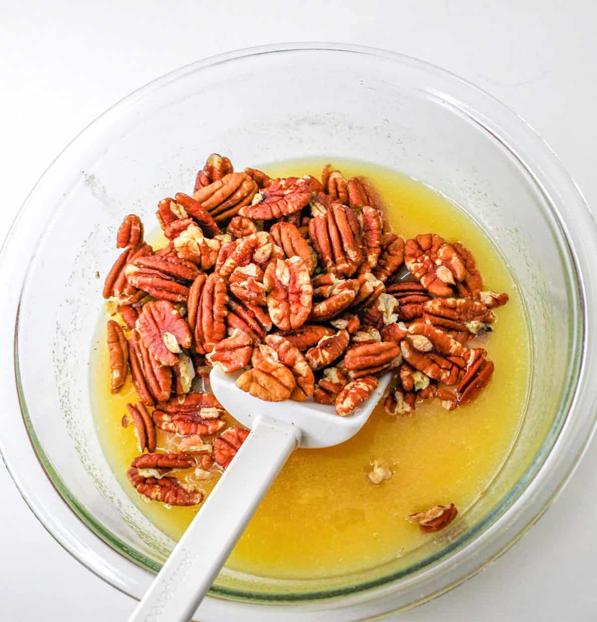 A clear bowl with pecan halves and a yellow liquid mixture, perfect for making Pumpkin Pie or Pecan Pie, being stirred with a white spatula on a white surface.