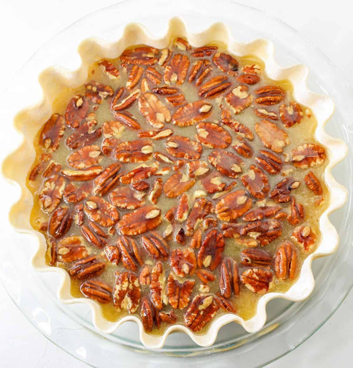 Unbaked Pumpkin Pecan Pie with a crimped pastry crust, filled with whole pecans and a glossy pumpkin-infused syrup mixture, in a glass pie dish.