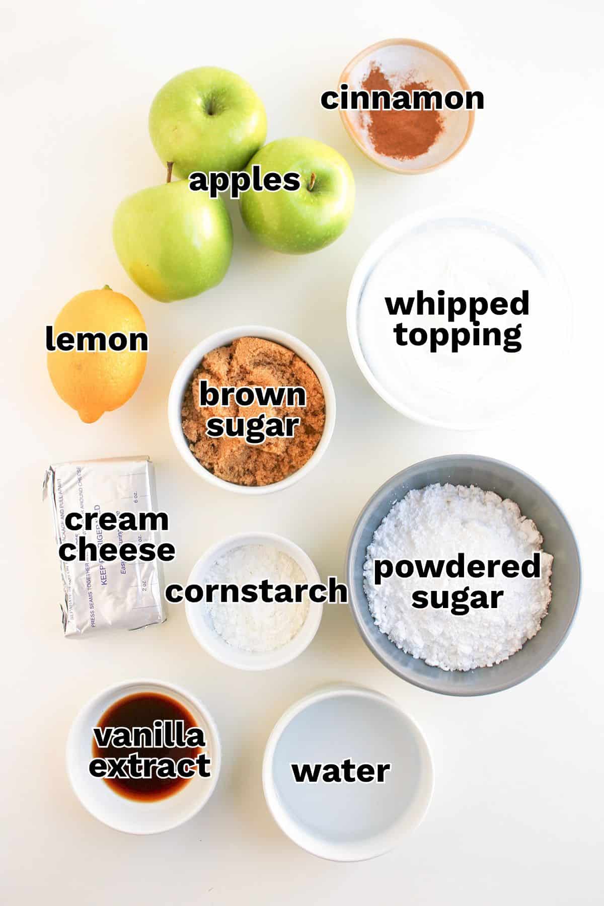 Top-down view of apples, lemon, brown sugar, powdered sugar, cornstarch, water, cream cheese, vanilla extract, whipped topping, and cinnamon arranged on a white surface—perfect for making a delicious Apple Pie Dip or a delightful apple dessert featuring creamy cream cheese.
