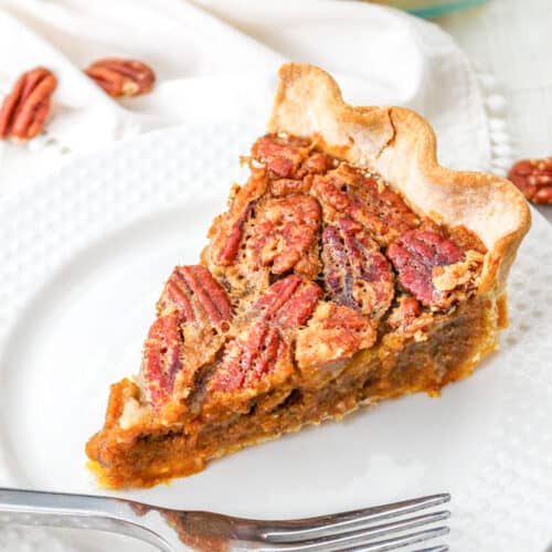 A slice of classic pecan pie sits on a white plate with a fork; the pie features a flaky crust and is topped with whole pecans, making it the perfect dessert for any pie lover.