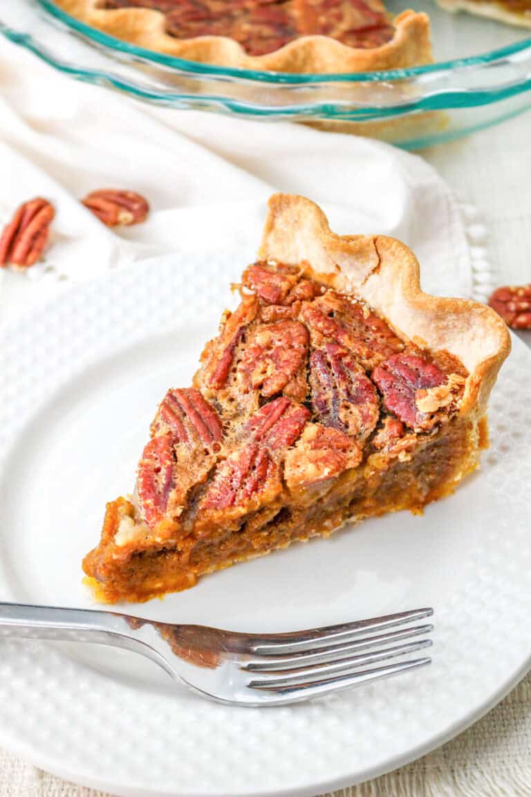 A slice of classic pecan pie sits on a white plate with a fork; the pie features a flaky crust and is topped with whole pecans, making it the perfect dessert for any pie lover.