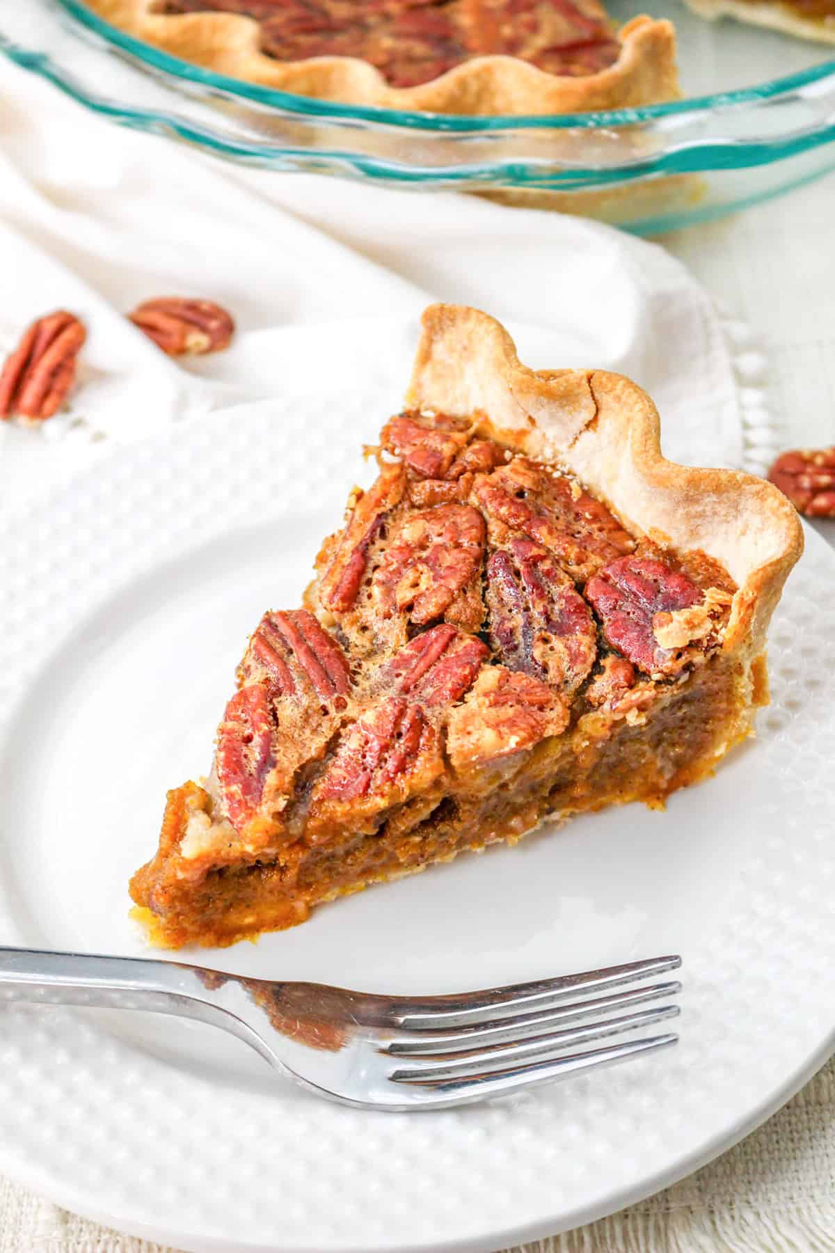 A slice of classic pecan pie sits on a white plate with a fork; the pie features a flaky crust and is topped with whole pecans, making it the perfect dessert for any pie lover.