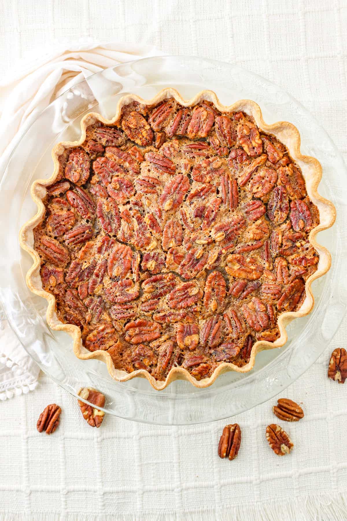 A baked pecan pie in a fluted crust sits on a clear glass pie dish, with a few pecan halves scattered on a white textured tablecloth, creating an inviting scene reminiscent of autumn gatherings and classic pumpkin pie desserts.