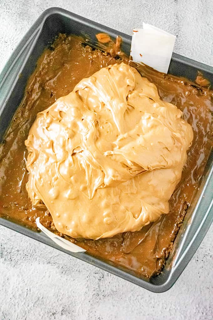 A baking pan filled with a dessert base, topped with a thick layer of creamy, light brown frosting or spread, reminiscent of indulgent Ferrero Rocher Cheesecake Bars.