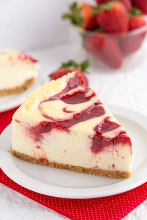 A slice of strawberry swirl cheesecake on a white plate, with a bowl of fresh strawberries in the background.