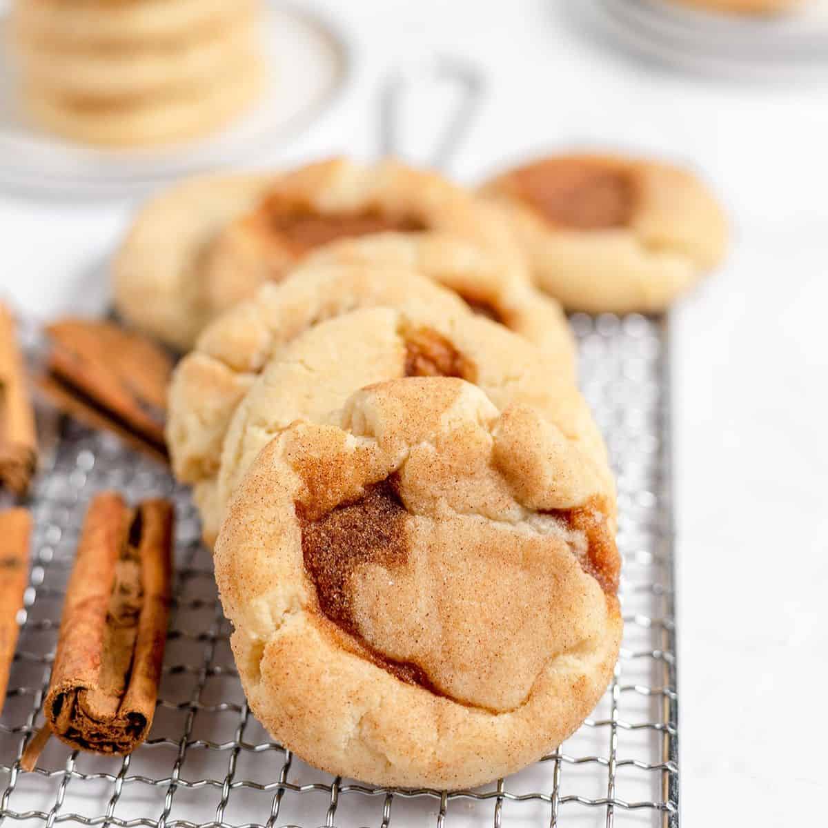 A row of cinnamon sugar snickerdoodles on a wire cooling rack with cinnamon sticks nearby.