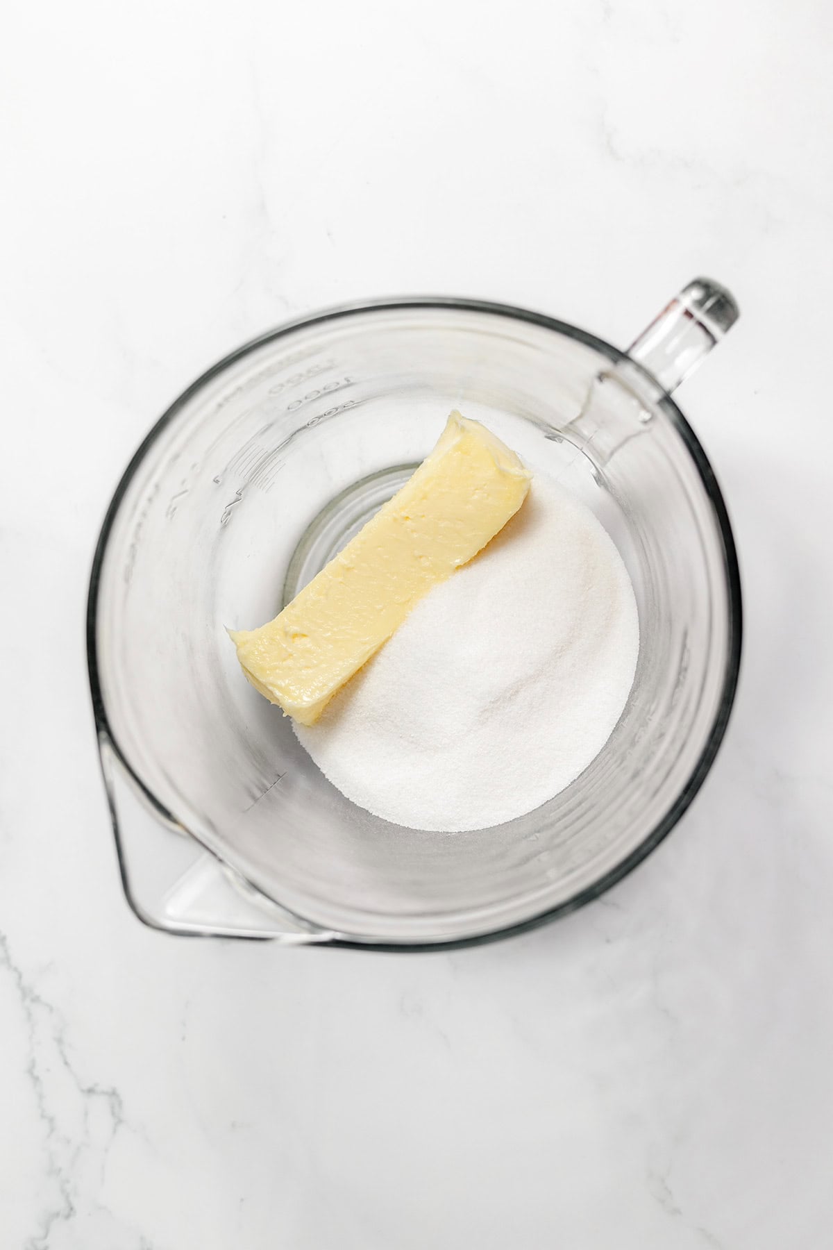 A stick of butter resting on a pile of granulated sugar inside a glass mixing bowl, perfect for making snickerdoodles.
