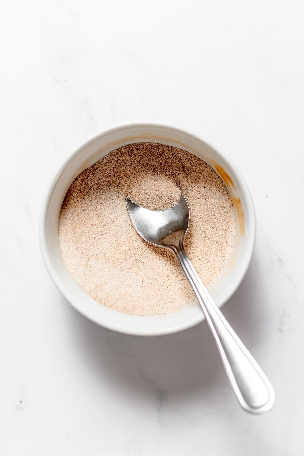 A white bowl filled with cinnamon sugar for snickerdoodle recipes, with a metal spoon inside.