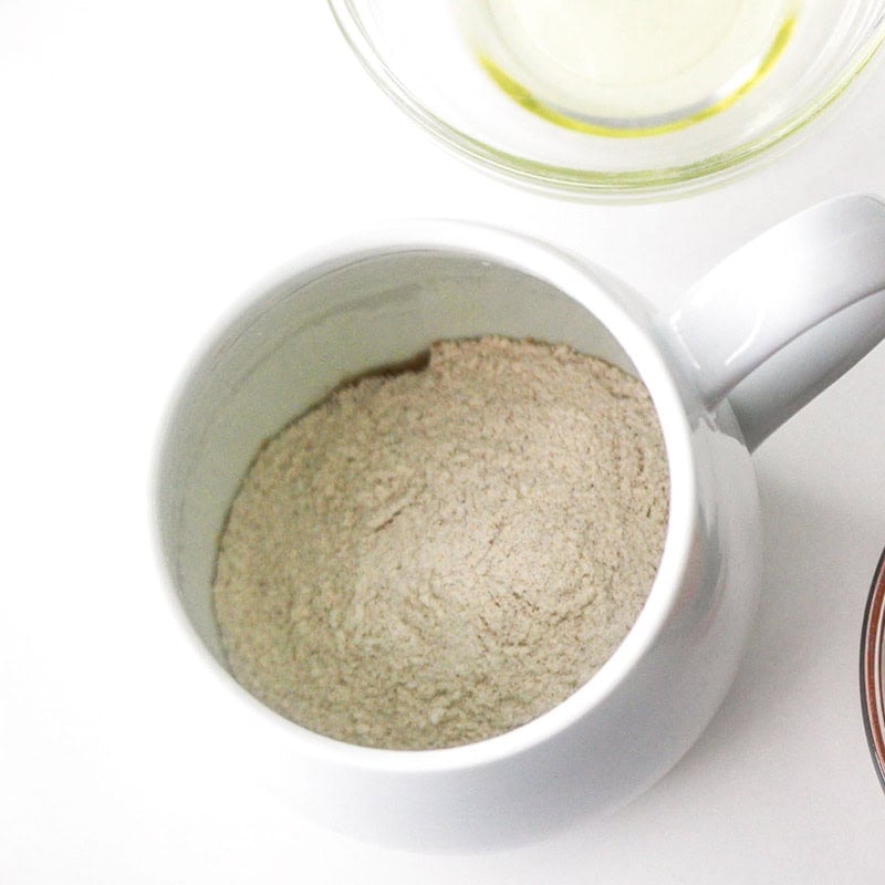 A white mug filled with beige powder for a pumpkin mug cake, next to a small glass bowl containing oil.