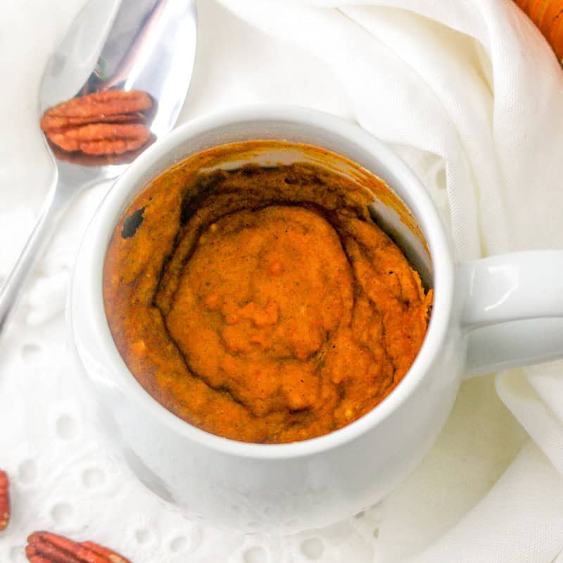 A white mug filled with pumpkin mug cake, served with pecans and a spoon on a white cloth.