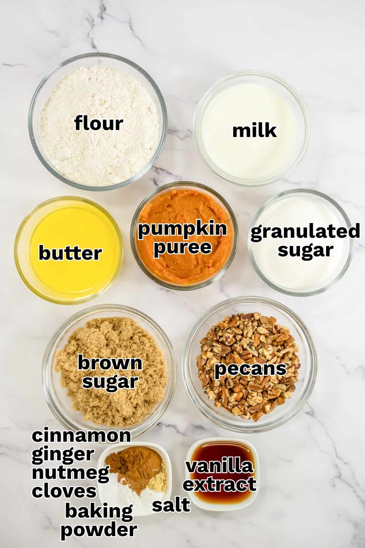 Glass bowls containing baking ingredients for a pumpkin pecan pudding: flour, milk, butter, pumpkin puree, granulated sugar, brown sugar, pecans, spices, baking powder, salt, and vanilla extract.