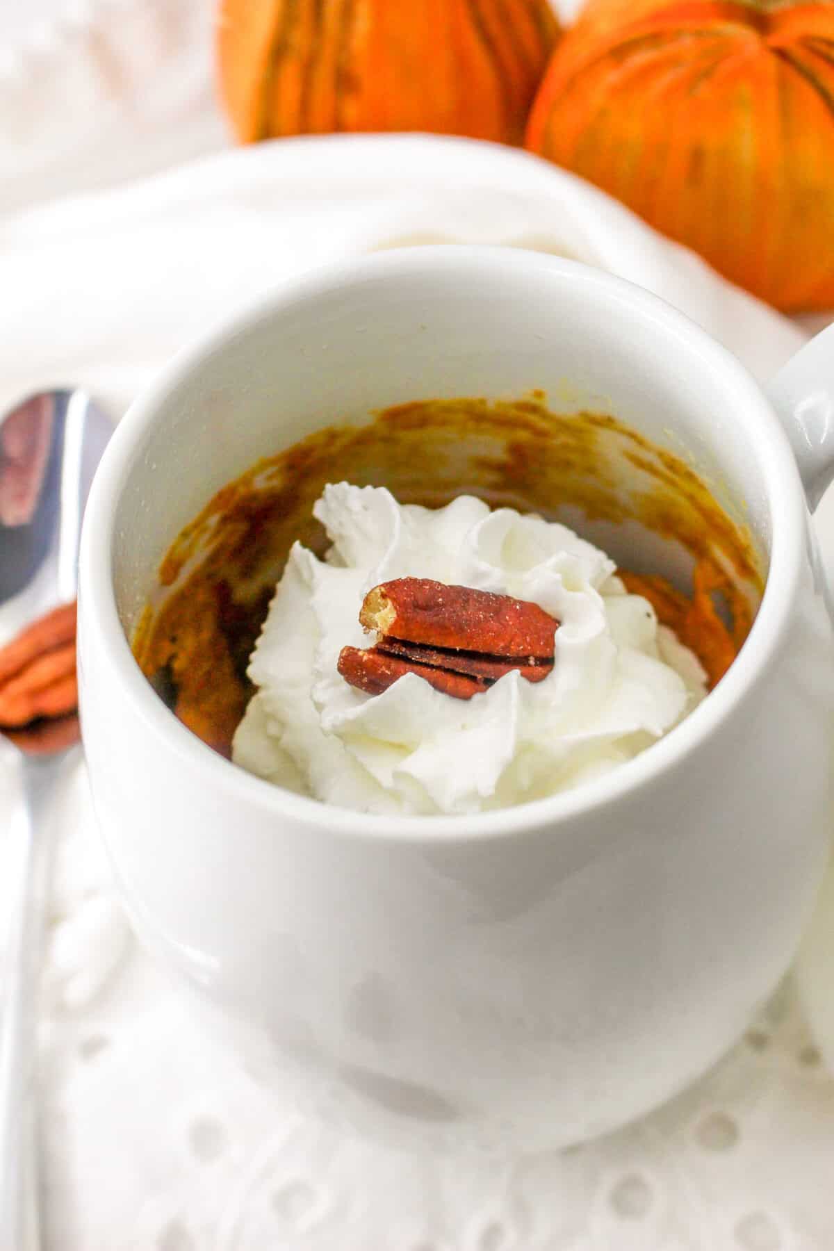 A pumpkin mug cake topped with whipped cream and a pecan, with mini pumpkins in the background.