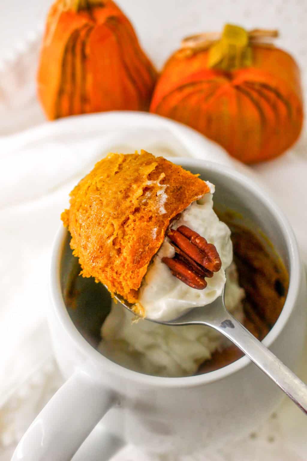A spoonful of pumpkin mug cake topped with whipped cream and a pecan, with two decorative pumpkins in the background.