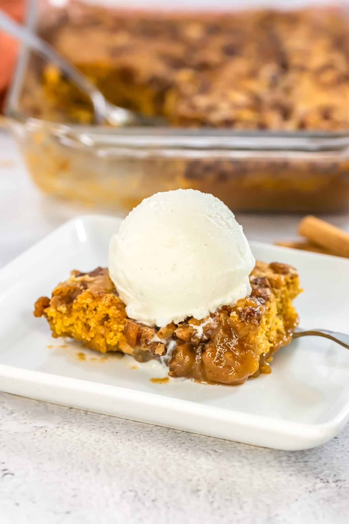 A square serving of pumpkin pecan dessert topped with a scoop of vanilla ice cream on a white plate.