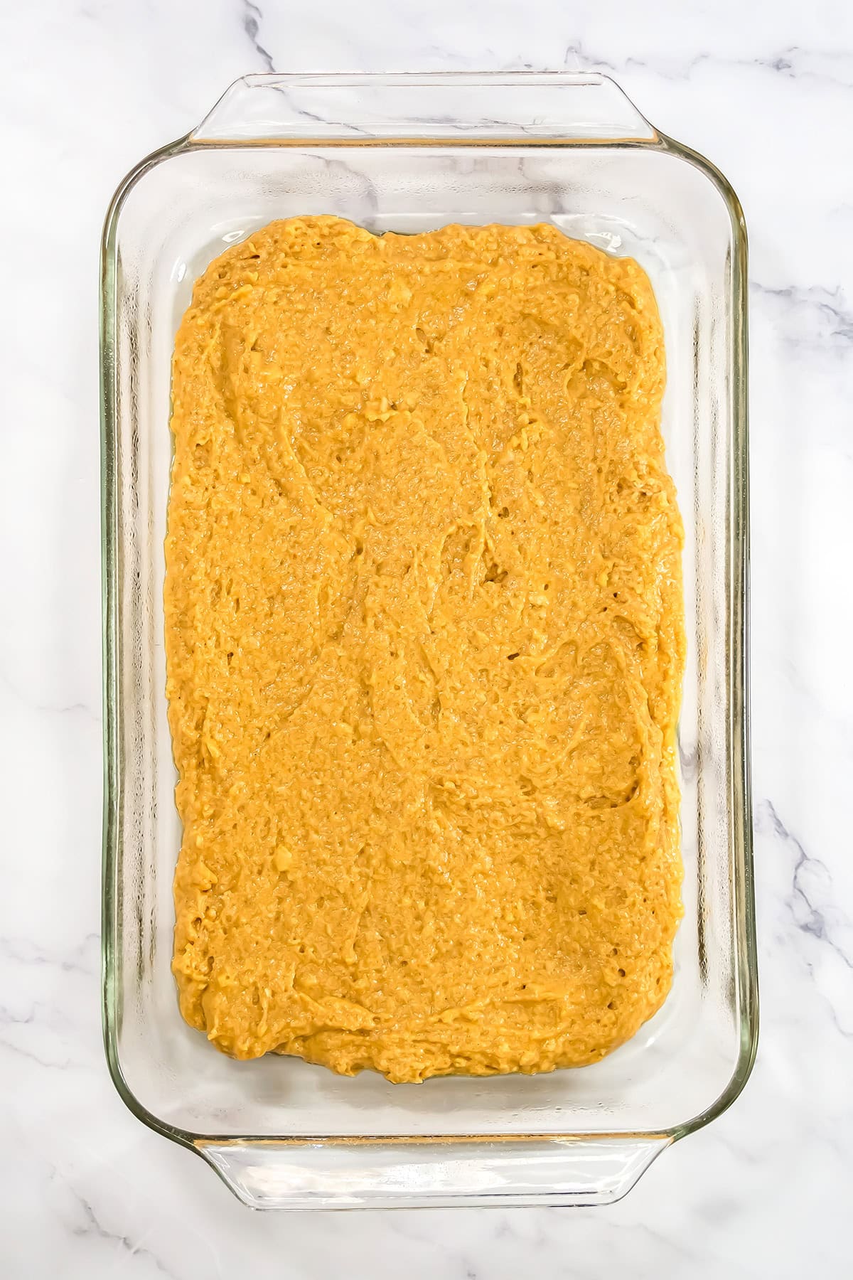 Raw pumpkin pudding cake batter spread evenly in a rectangular glass baking dish on a marble surface.